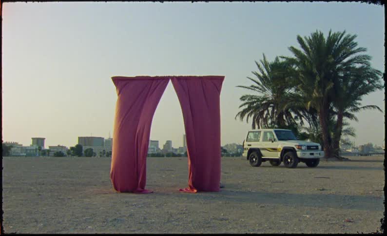 A red fabric archway stands in an open sandy area with a white offroad vehicle parked nearby and palm trees and city buildings visible in the background