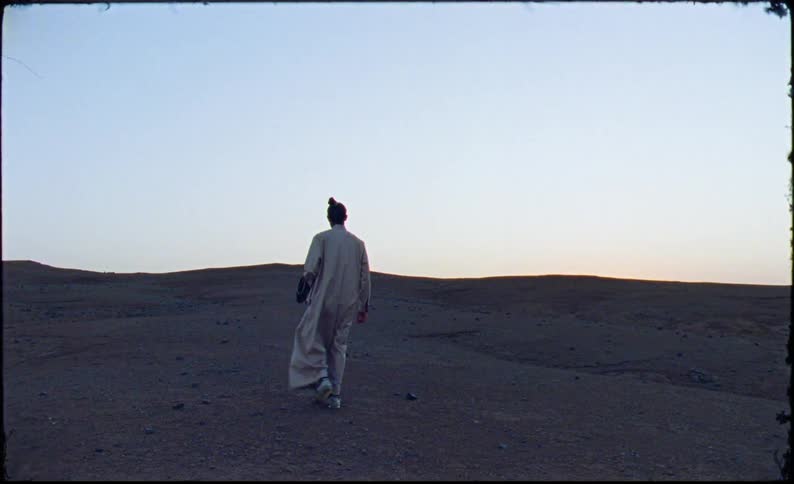 A solitary figure wearing a long lightcolored garment walks across a barren hilly landscape at dusk holding an object in one hand silhouetted against a clear sky