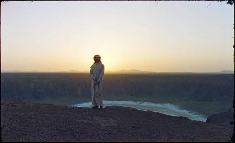 A man in traditional attire stands on a cliff overlooking a scenic sunset