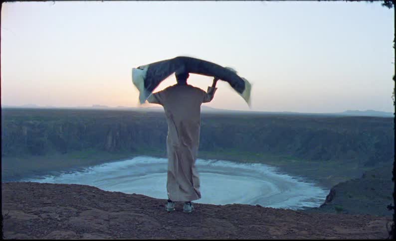 A man is holding a loose piece of clothing that is flying in the wind as he stands on the edge of a high hill looking at the horizon