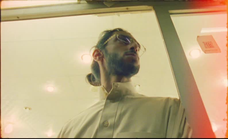 A man with tied hair and wearing glasses is leaning against the door of a store that is all lit up inside