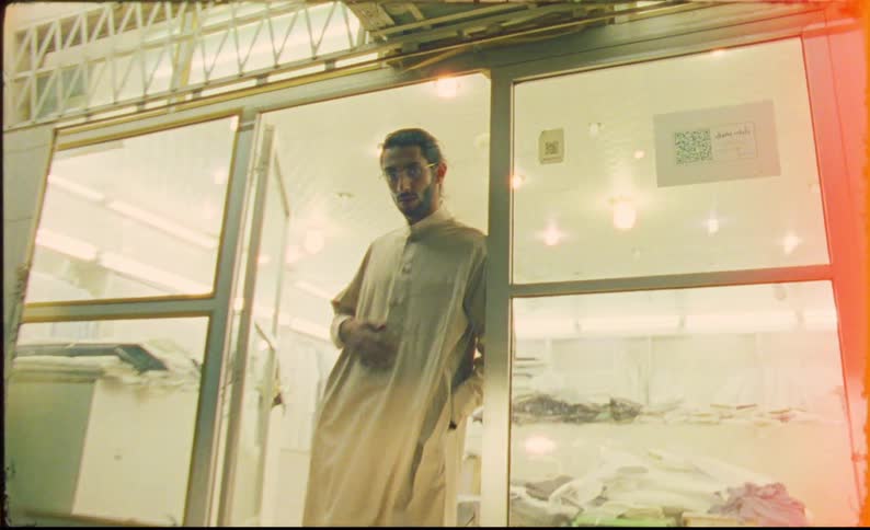 A man wearing a traditional thawb standing pensively outside a brightly lit storefront with glass doors while air conditioning units are mounted above and the ambiance suggests a quiet urban setting
