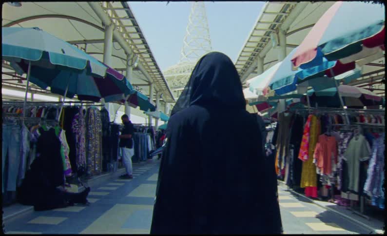 A woman in a black burka is walking through an outdoor market with various stalls selling clothing under colorful canopies