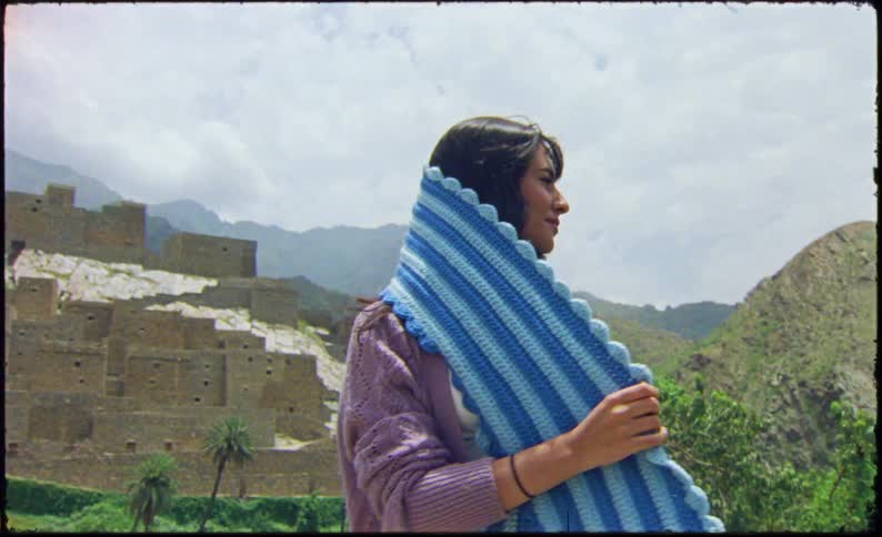 A person wearing a purple sweater and a blue and white striped scarf stands in front of an ancient fortress in the mountains