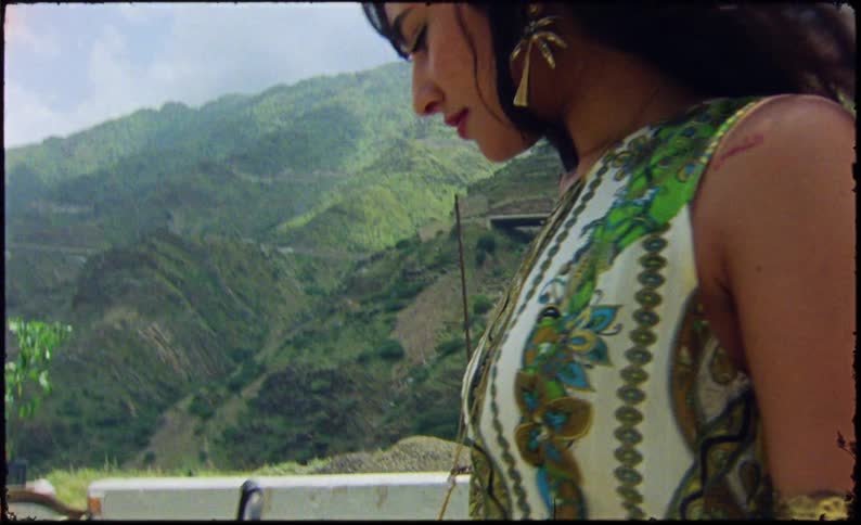 A woman in a green dress walking to the left and behind her there are mountains covered in green vegetation