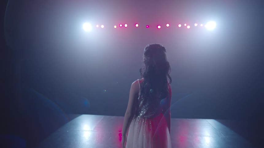 A performer stands on a stage arms raised towards glowing lights creating a dramatic and ethereal scene