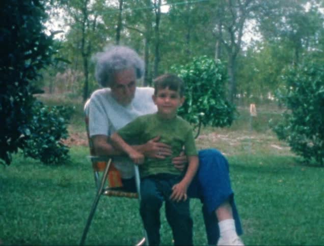 A lady is sitting on a chair in a yard with many trees and she has her grandson on her lap and he is looking straight ahead