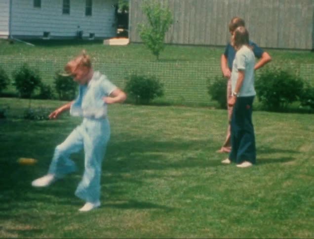 S a child playing an ankle skip game in a backyard swinging the toy around one ankle while jumping over it two adults and another child stand nearby watching the activity in the grassy area surrounded by a fence and houses