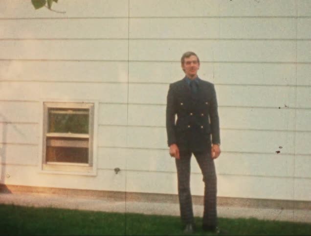 A man standing wearing formal clothing and behind him there is a white house with small windows and in an old image