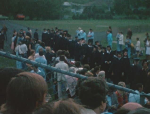Some people are sitting on a separate stand with a metal fence and there is a lawn in front with many people standing and some of them are wearing black clothes