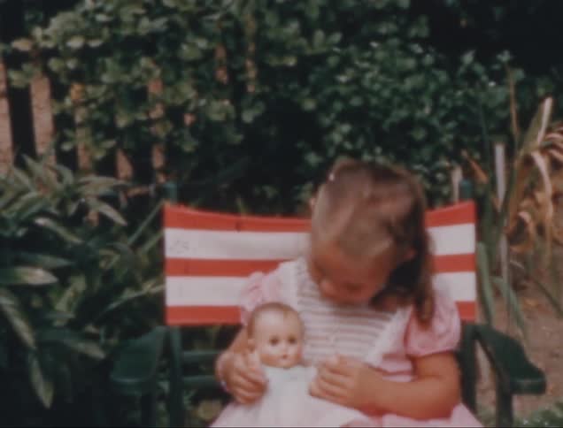 A little blonde girl is sitting on a red and white chair and is playing with her doll