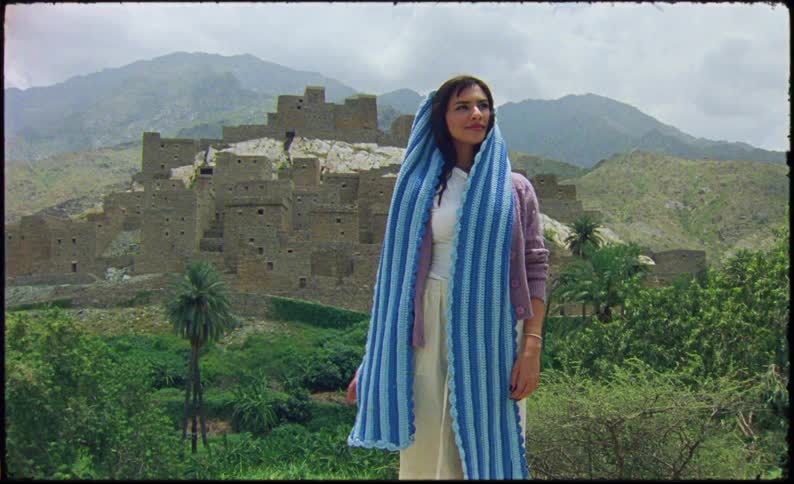 A woman standing with a subtle smile is adjusting a blue piece of clothing around her neck and behind her there are some old buildings