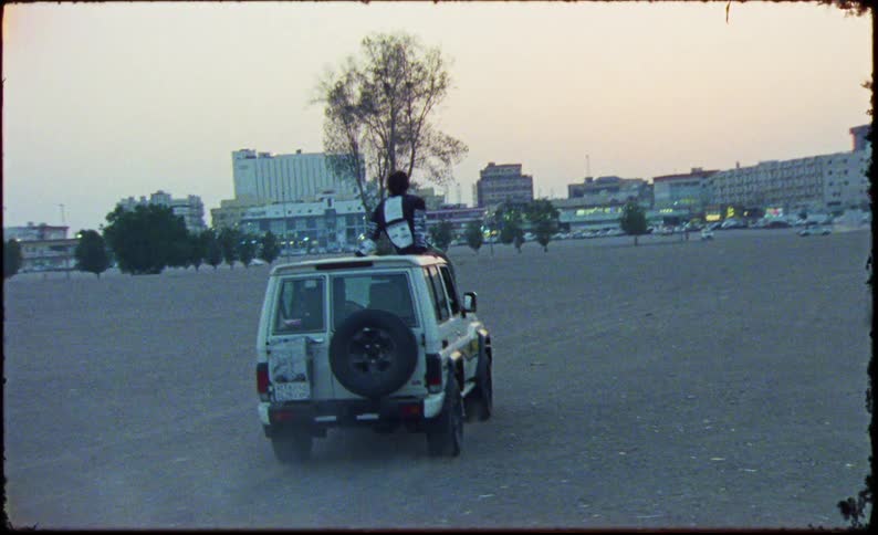 Man on top of a jeep moving through a desert area with a city in the background