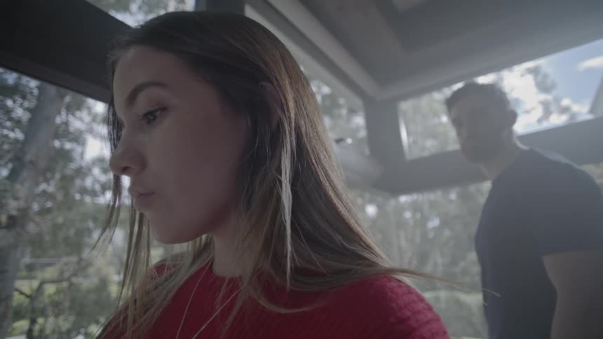 A woman in a red sweater sits indoors near large windows with a view of trees while a man in a dark shirt stands behind her