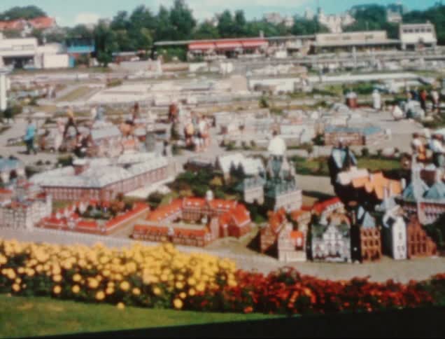 A detailed miniature model of a cityscape includes various buildings roads and greenery with vibrant yellow and red flowers in the foreground