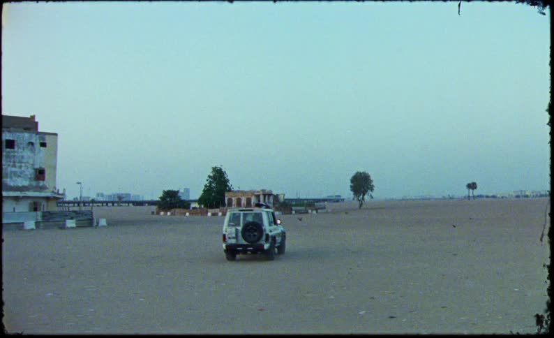 A white suv is driving across a vast empty desertlike area with a man sitting on its roof