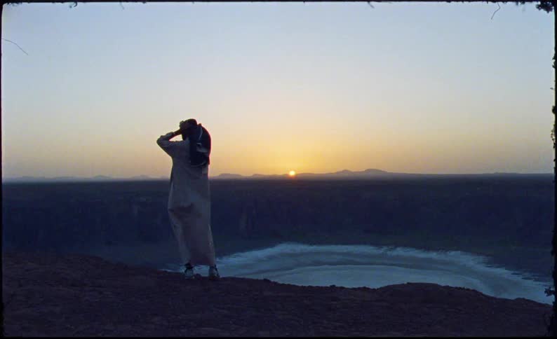 Person in arabic clothing on top of a mountain with a lake in the background and the sun rising on the horizon