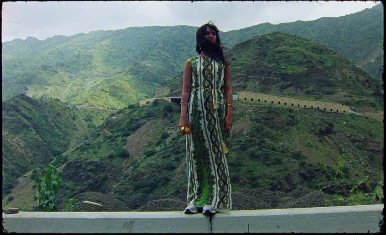 Woman in a long dress holding a fruit on top of a concrete wall with a mountain valley in the background