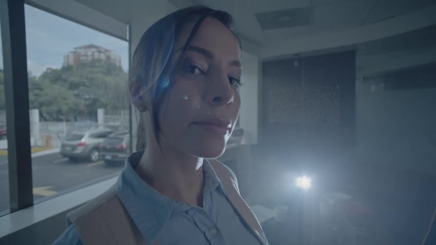 A person is standing indoors near a large window with a view of a parking lot while a bright light shines from behind