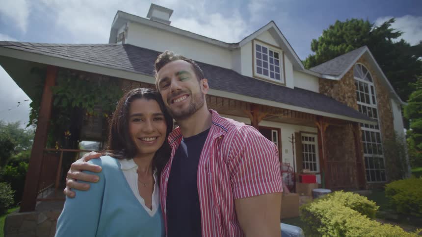 A man and a woman are hugging and smiling as they look forward and behind them is a house with several windows