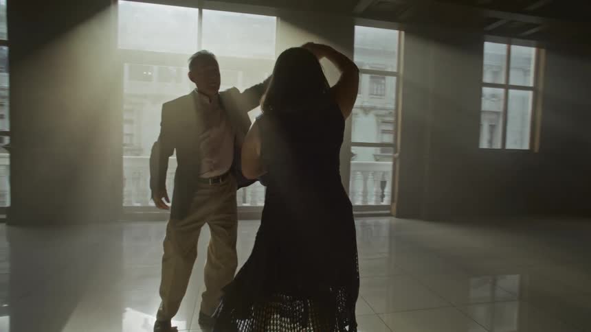A man and a woman dancing in an empty room with several windows and the man spins the woman around and her skirt moves