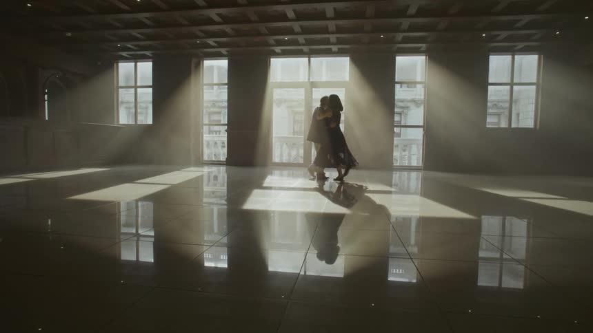 A couple is dancing in a large empty room with sunlight streaming through tall windows casting long shadows and reflections on the polished floor