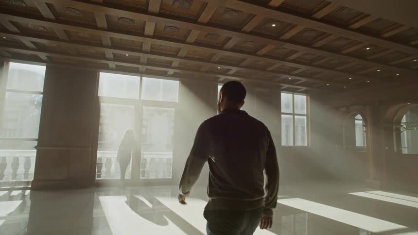 Man in a lounge walking towards a woman standing at the window