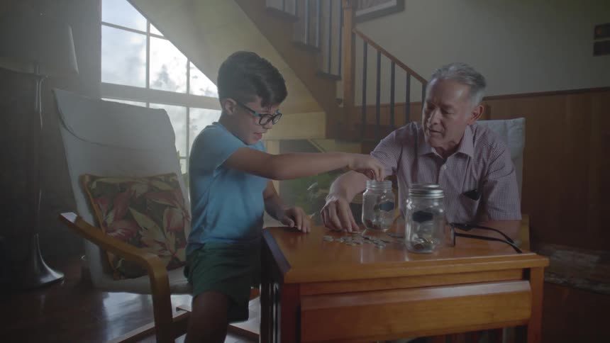 A little boy and an elderly man are sitting and putting coins that are on a table into glass jars
