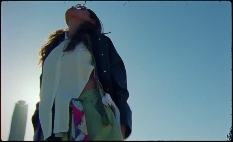 Woman models clothing while standing on front of a concrete barrier with the city behind her