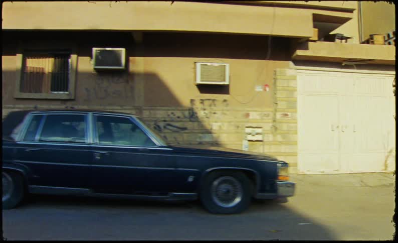A vintage cadillac sedan is parked on a street lined with brick buildings bathed in the warm light of the setting sun