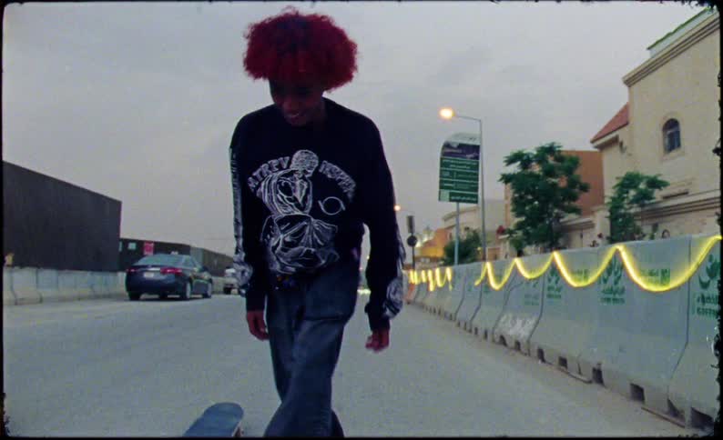 A skateboarder midtrick on an urban street wearing a graphic longsleeve shirt and loose pants with cars and construction cranes visible in the background