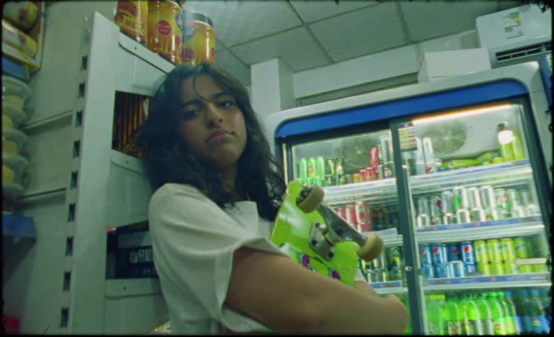 A girl with dark wavy hair a white shirt and gray shorts stands in front of a food shelf holding a green skateboard in the supermarket