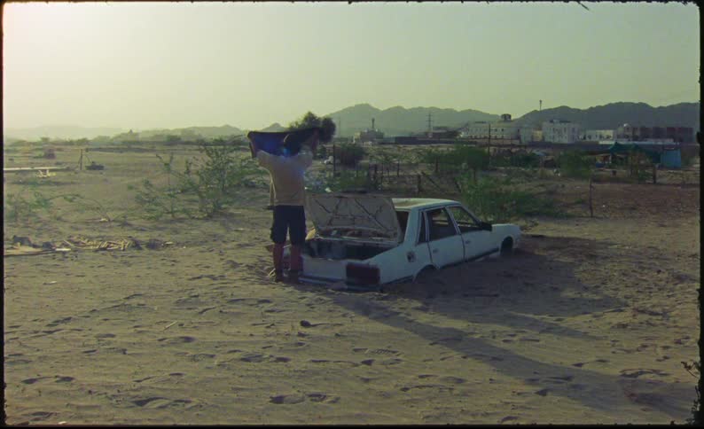 A person examines the trunk of a car buried in a sandy desert