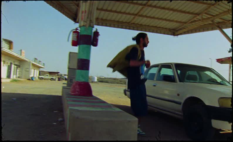 A bearded man in a brown vest black tshirt and shorts sits on a car hood at a gas station