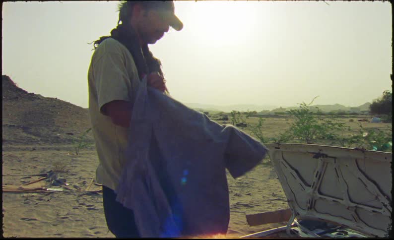 Man looking at a blue shirt and putting it on while standing in a desert environment with sparse vegetation