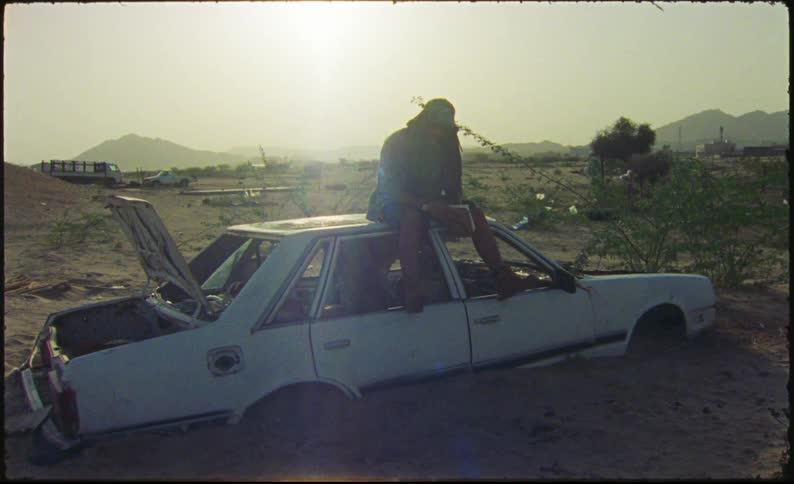 Man sitting on top of an old white car buried up to its wheel wells in the sand