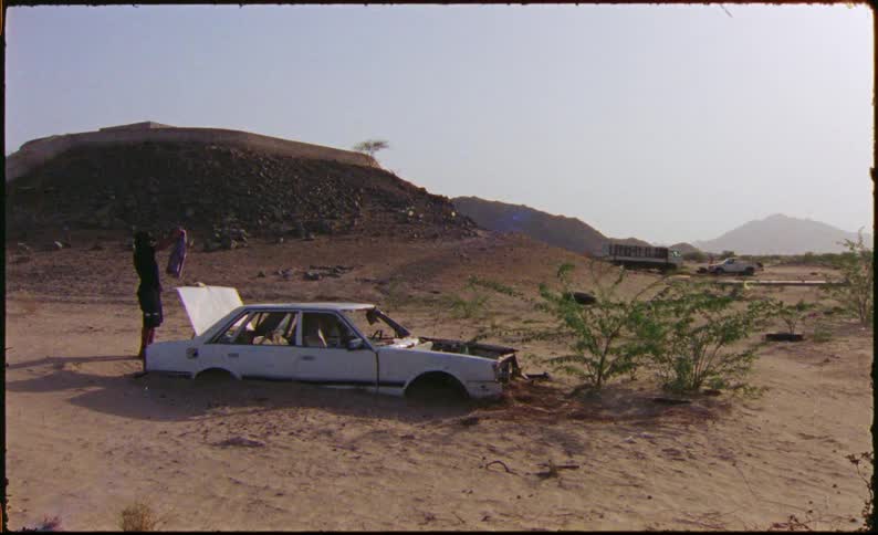 A wrecked car partially buried in sand in a desert landscape with a person holding up a shirt