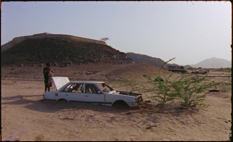 A man inspects the hood of an abandoned car and lifts up a t shirt to sway in the wind in a desert landscape
