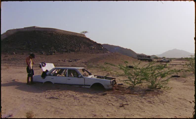 Man stands near a ruined abandoned car and changes his clothes with hills in the background