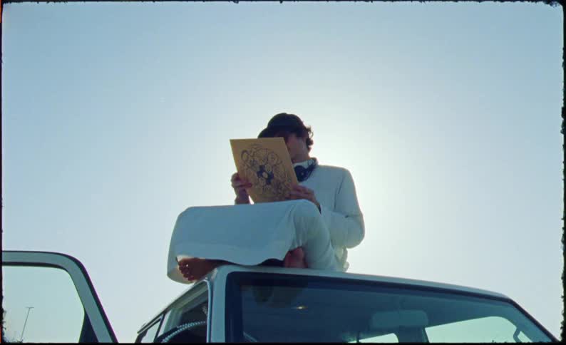 Man in cap and glasses sitting on the roof of a car watching a vinyl record