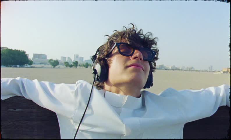 A man wearing a white shirt sunglasses and headphones leaning back with arms outstretched seemingly enjoying a serene moment outdoors with an urban skyline in the background traveling by boat