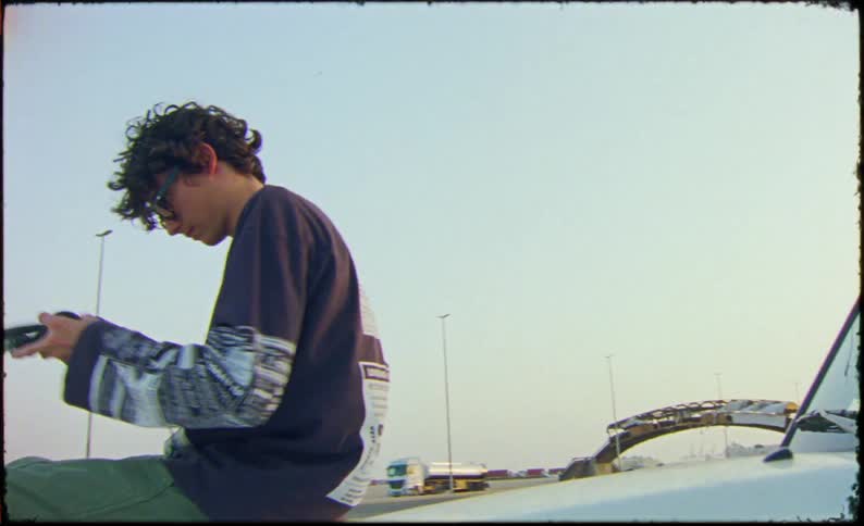 Teen is listening to headphones and sitting on a parked car by the highway