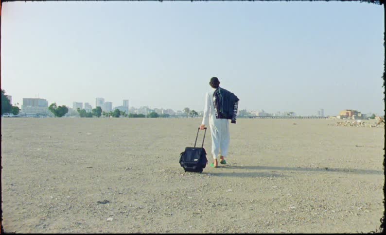 A person in loose white clothing stands with a wheeled suitcase next to him in a desert and then turns around and starts walking away