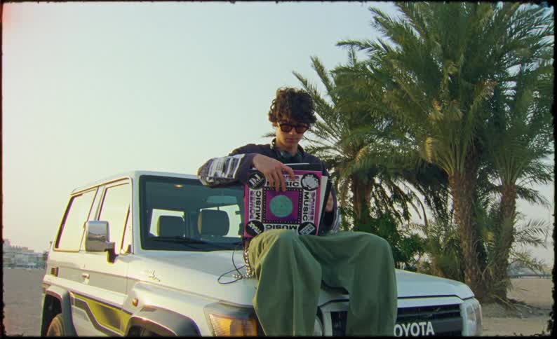 Man wearing sunglasses sitting on the hood of a vehicle watching music records