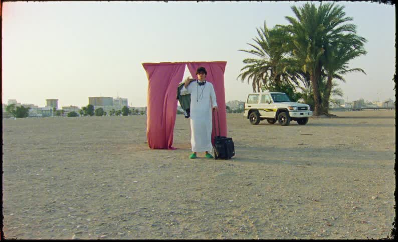 Man pulls a suitcase and carries clothes to pose next to curtains on a frame with the city in the background
