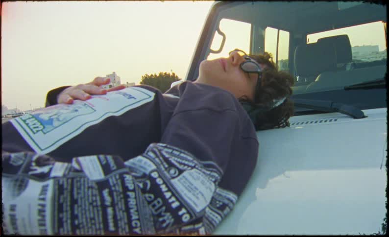 Man smiles and listens to music while lying on the hood of his car while the sun shines from the horizon