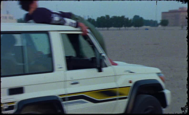Man on top of a white car moving through a desert area with buildings in the background