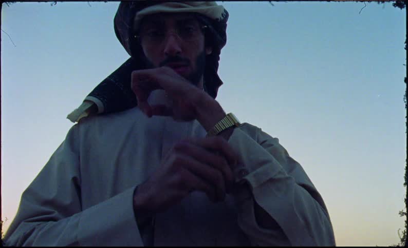 A young man wearing traditional middle eastern attire stands on the edge of a large crater at sunset