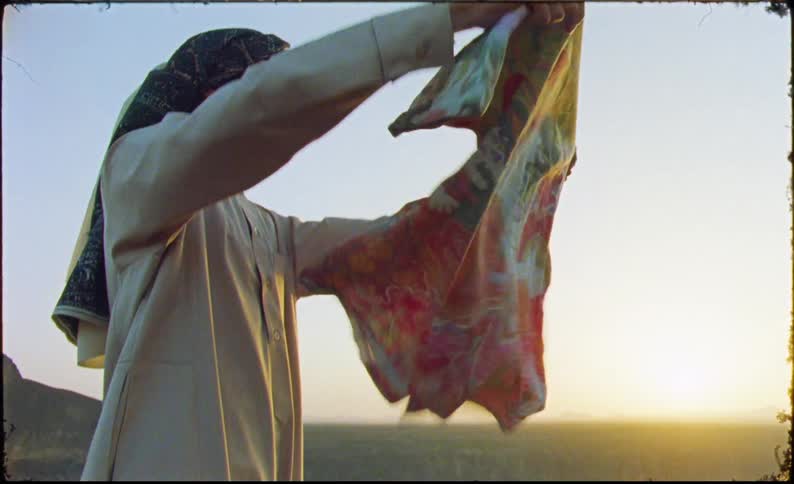 Man reaching for a handkerchief inside a suitcase while standing in a natural area with the sun on the horizon
