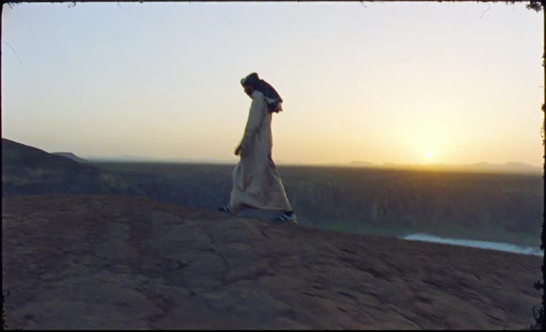 Man in an arab robe walking up a hill with the sun on the horizon